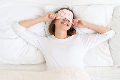 Young female waking in morning on white pillows and sheets, stretching elbows, wearing sleeping mask on eyes, enjoying late morning in bed